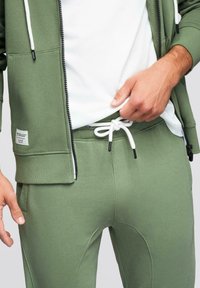 Homme portant un sweat à capuche zippé vert olive et un pantalon de survêtement assorti avec un cordon blanc, ajustant la taille au-dessus d'un T-shirt blanc.