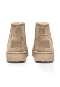 Beżowe zamszowe botki za kostkę z teksturowanym ściegiem po bokach, gumową podeszwą oraz wytłoczonym logo z tyłu. Prosty, funkcjonalny design.