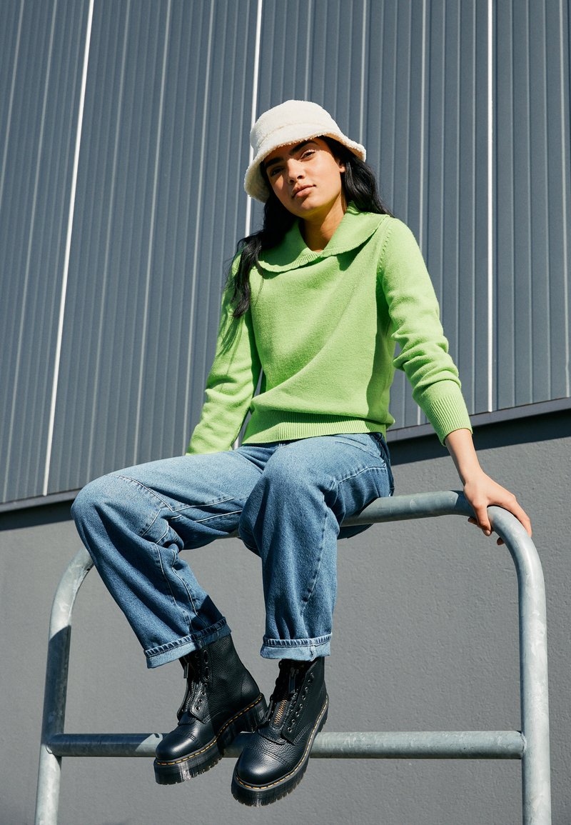 Green collared sweater, relaxed blue jeans, and black lace-up boots. Model sitting on a metal railing against a gray backdrop.