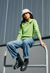 Pull vert avec un large col, jeans baggy bleu clair, et bottines noires à semelle épaisse, assis sur une rambarde en métal.