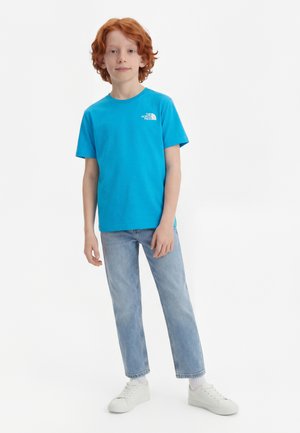 Ragazzo giovane dai capelli rossi che indossa una maglietta blu di The North Face, jeans azzurri, calzini bianchi e scarpe da ginnastica bianche, in piedi contro uno sfondo bianco.