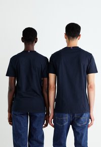 To personer iført identiske marineblå T-shirts, side om side, med et enkelt design og rund hals. T-shirtsene har en lille stribet detalje ved halsen.