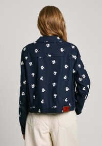 Mörkblå kort jacka i tyg med vit blommig brodyr, knäppningar vid ärmsluten och en läderpatch-märke vid nederkanten på baksidan.