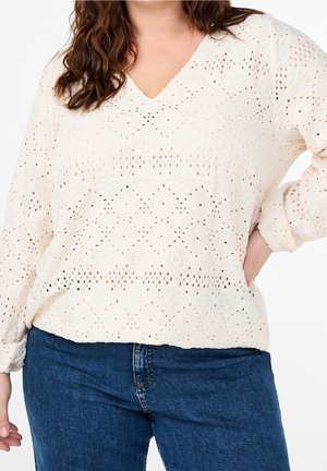 Femme portant un chemisier crème en dentelle à manches longues bouffantes et col en V, avec un jean bleu, debout, une main sur la hanche.