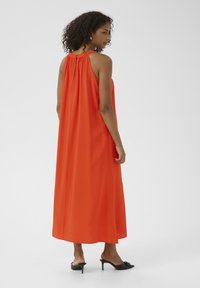 Abito maxi arancione senza maniche con colletto arricciato, silhouette fluida e tessuto leggero. Indossato con mules neri con tacco.