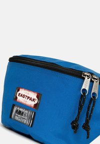 Pochette cosmetica in tessuto blu con doppie chiusure a zip, dettagli neri e etichette Eastpak e MM6 sul lato. Superficie testurizzata.