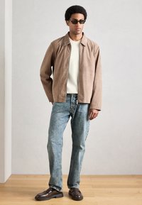 Barna velúr dzseki krémszínű pulóver felett, világoskék farmerrel és sötétbarna mokaszinnal párosítva. A fekete napszemüveg teszi teljessé a megjelenést.