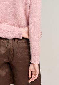 Hellrosa, gestrickter Pullover mit gerippten Bündchen, kombiniert mit braunen, strukturierten Shorts. Der Stoff des Pullovers wirkt weich und flauschig.