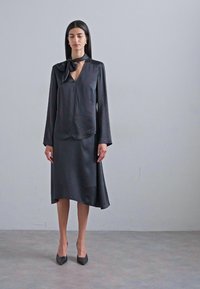 Svart satengbluse med knyte-detalj og dyp V-hals, kombinert med et kne-lang skjørt. Begge har en glatt tekstur og flytende design.