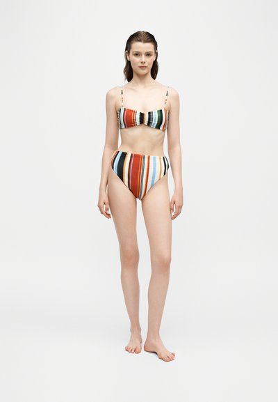 Femme debout pieds nus portant un bikini multicolore à rayures verticales avec un bas taille haute sur un fond blanc uni.