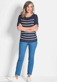 Navy-blauer, gestreifter Strickpullover mit weißen horizontalen Streifen, kombiniert mit hellblauen Jeans mit umgeschlagenem Saum und weißen Sneakers. Das Model steht mit verschränkten Armen.