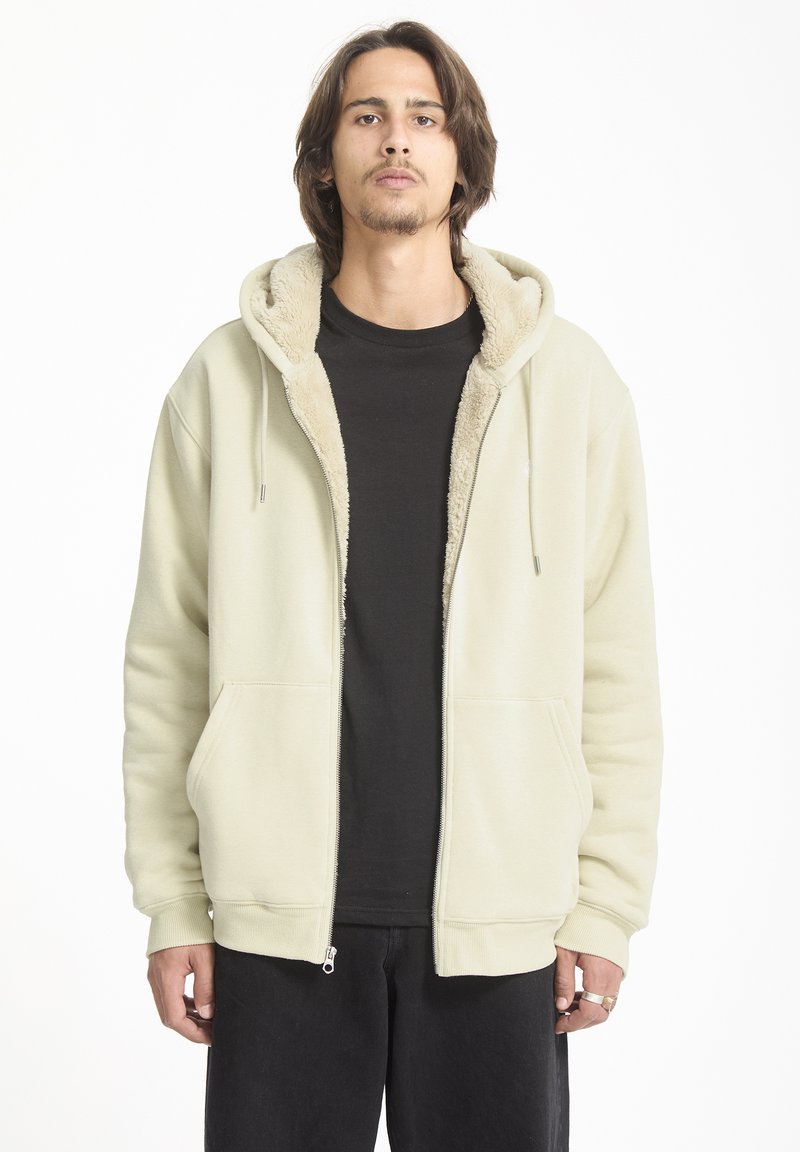Sudadera con cremallera de un suave color beige claro, con interior forrado de piel, bolsillos tipo canguro y puños acanalados, usada sobre una camiseta negra.