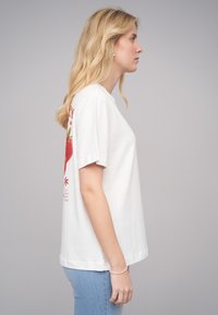 T-shirt en coton blanc à manches courtes, avec un grand graphique de fraise rouge au dos et du texte. Coupe décontractée et encolure standard.