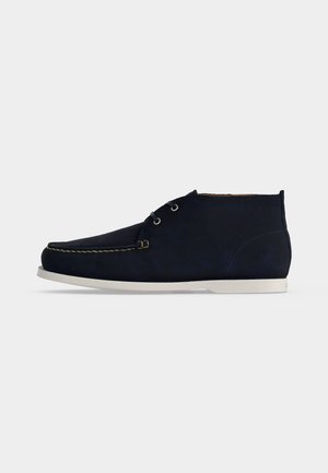 Bottines en suède bleu marine pour hommes, avec semelles blanches et fermeture à lacets sur fond clair uni.