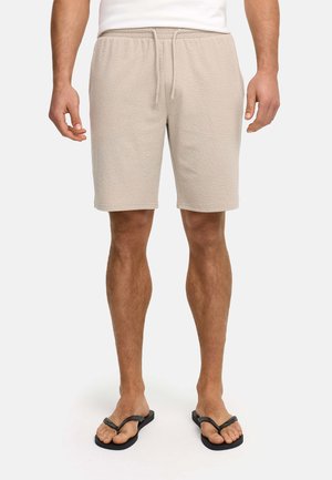 Mann trägt beige, strukturierte Shorts mit Kordelzug, weißes Hemd und schwarze Flip-Flops, steht vor einem einfarbigen hellen Hintergrund.