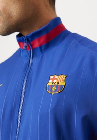 Modrá športová bunda s červeným pruhom na golieri, s pásmi a zlatým logom FC Barcelona na ľavej hrudi. Zipsový predok.