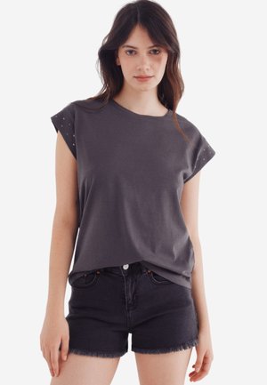 Graues T-Shirt mit kurzen Ärmeln und lockerer Passform, mit Nietenakzenten an den Schultern und einer glatten Textur. Kombiniert mit schwarzen Denim-Shorts.