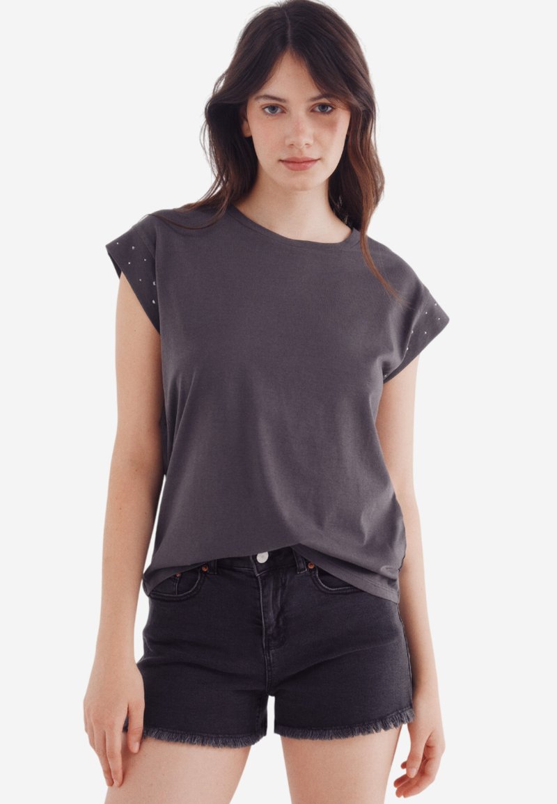Grå kortærmet t-shirt med en afslappet pasform, der har studs detaljer på skuldrene og en glat tekstur. Sammen med sorte denim shorts.