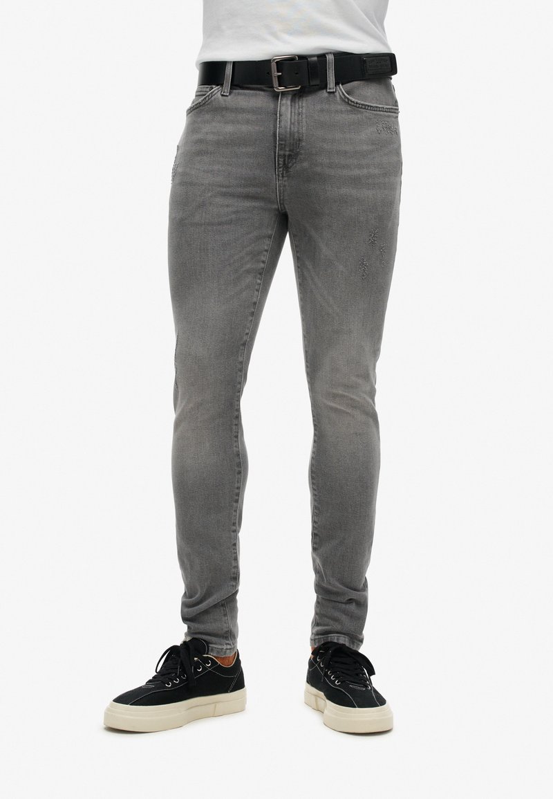 Jeans gris ajustados con detalles desgastados, cinturón negro y zapatillas casuales negras con suelas crema y cordones blancos, mostrando un look moderno.