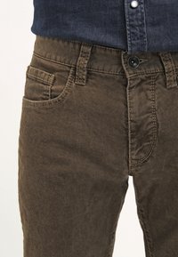 Nærbillede af brune corduroy-bukser med knap- og lynlås-lukning, båret med en mørkeblå skjorte, der er stoppet ind.
