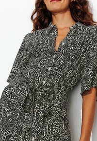 Robe imprimée paisley noir et blanc avec un col, des manches courtes, un devant à boutons et un détail de taille nouée. Tissu doux et texturé.