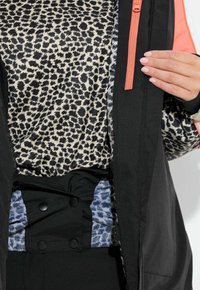 Černá bunda s oranžovým zipem přes béžový top s leopardím potiskem. Má černý pasový lem s modrými vzory a dvojími knoflíky.