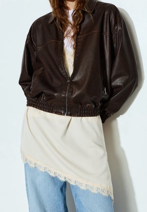 Personne portant une veste bomber en cuir marron foncé par-dessus une robe nuisette crème bordée de dentelle, associée à un jean bleu clair, debout contre un mur blanc.