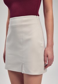 Ljus beige mini kjol med en mjuk textur, försedd med en framklippning och en diskret siddragkedja. Matchas med en åtsittande vinröd ribbad topp.