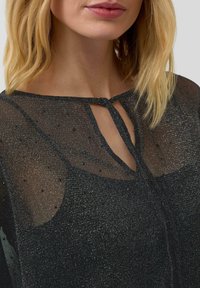 Vrouw met schouderlang blond haar, gekleed in een zwarte glinsterende sheer top met een keyhole halslijnen en een klein gestippeld patroon.