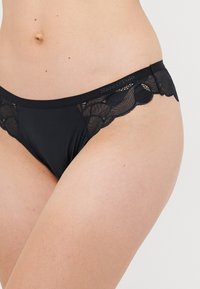 Mutande in pizzo nero in stile bikini sui fianchi e sulla parte superiore delle cosce di una persona, con il logo Marc O’Polo sulla cintura.