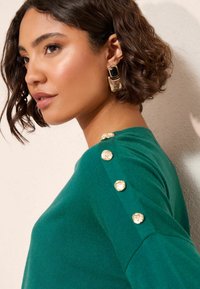 Zielony sweter z fakturowaną powierzchnią i złotymi guzikami na ramieniu. Projekt podkreśla dopasowany krój i miękką tkaninę.