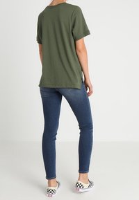 Olivgrön bomullströja med uppvikta ärmar, tillsammans med mörkblå skinny jeans och svart-vita rutiga slip-on skor.