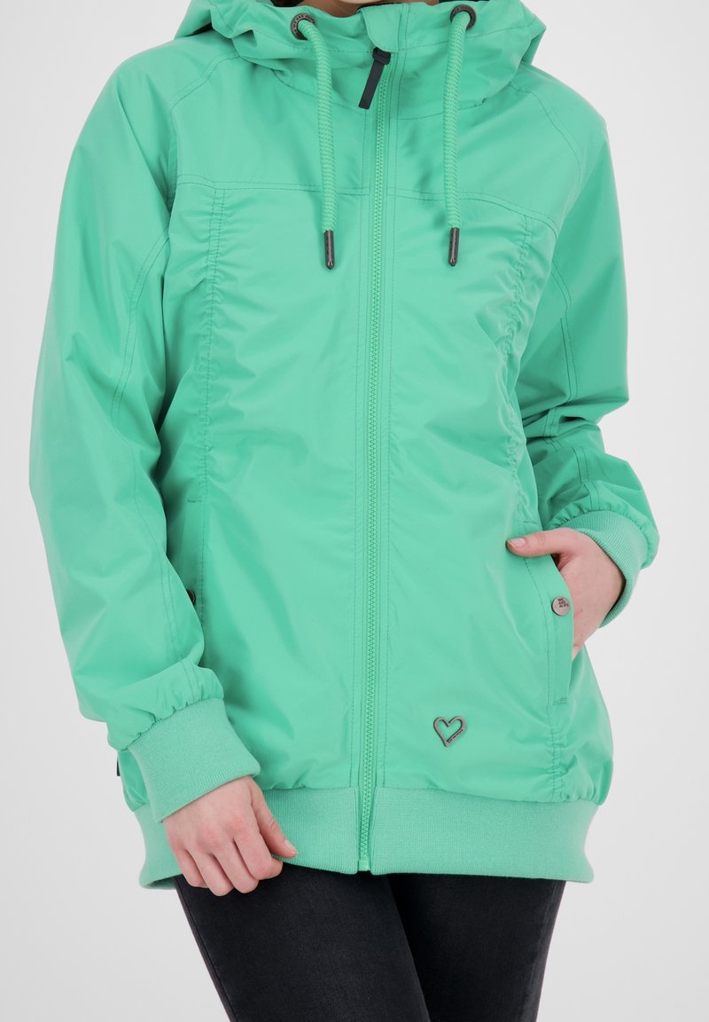 Personne portant une veste à capuche zippée vert menthe avec cordons, poignets et ourlet côtelés, une main dans la poche latérale, associée à un pantalon noir.