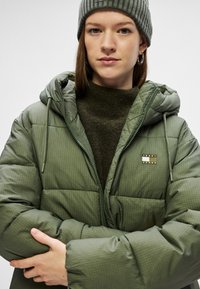 Giacca puffer verde oliva con cappuccio, design trapuntato, zip frontale e patch con logo. Indossata sopra un maglione scuro e un cappello di lana.