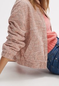 Teksturowany, lekki różowy kardigan z podwiniętymi rękawami, o luźnym kroju. Noszony na jasnoróżowej bluzce i niebieskich dżinsach.