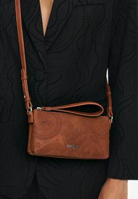 Sac en cuir marron à bandoulière avec un motif floral embossé, une fermeture éclair et une sangle ajustable, présenté sur un tissu noir texturé.