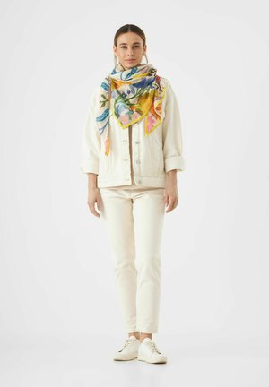 Veste en denim blanche sur un haut beige, assortie à un pantalon clair et des baskets blanches. Un foulard floral coloré ajoute des accents vivants.