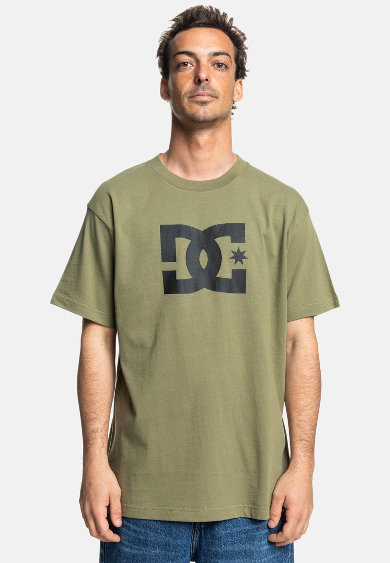 Camiseta de algodón verde oliva con mangas cortas, que presenta un gran logotipo gráfico negro en el centro. Corte relajado con un cuello redondo.