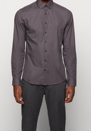 Mężczyzna w dopasowanej, długorękawowej koszuli na guziki z drobnym wzorem w kratkę oraz ciemnoszarych spodniach.