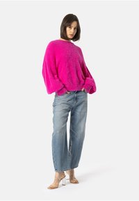 Pūkains rozā adīts džemperis ar nokritušām plecu daļām, kombinēts ar gaiši ziliem nodilušiem džinsiem un caurspīdīgām papēžu sandalēm. Vienkārša, brīva silueta.