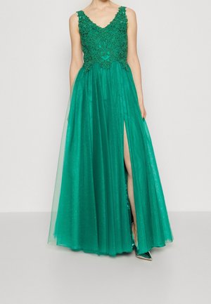 Robe verte sans manches avec corsage en dentelle, jupe en tulle scintillante et fente haute dévoilant une jambe et une chaussure pointue métallique.