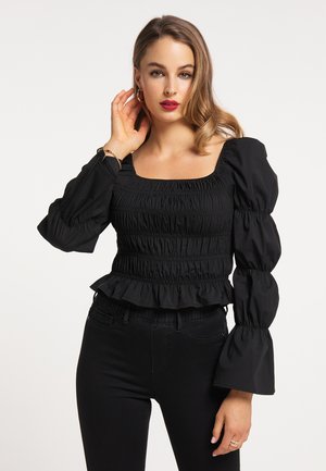 Junge Frau mit welligem Haar, die ein eng anliegendes schwarzes Oberteil mit Puffärmeln und schwarze Hosen trägt und mit der Hand am Gesicht posiert.