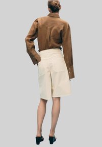Chemise marron à manches longues et short crème. La chemise possède des poignets à bouton, tandis que le short a une taille haute et deux poches arrière. Sandales à talons noires.