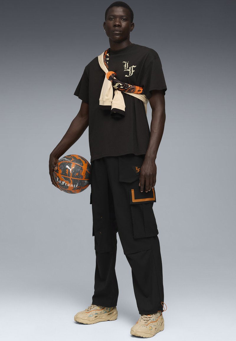 Sort t-shirt med logo, beige shorts om halsen, sorte cargo bukser med orange detaljer, holder en mønstret basketball.