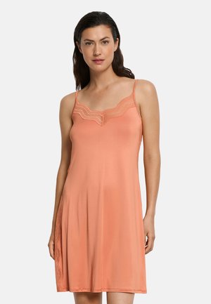 Kvinde iført en koralfarvet ærmeløs slip-kjole med blondekant på udskæringen og siderne, stående mod en ensartet lys baggrund.