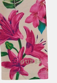 Tessuto con stampa floreale che presenta grandi gigli rosa e accenti di foglie verdi su uno sfondo beige. "GUESS" ricamato in oro nella parte inferiore.