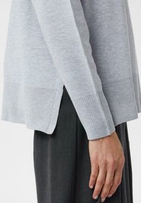 Nahaufnahme einer Person, die einen hellgrauen gerippten Pullover mit seitlichem Schlitz und dunkelgrauen Hosen trägt, der Arm entspannt an ihrer Seite.
