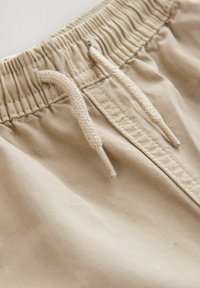 Pantaloni corti beige con vita elastica e tessuto arricciato, dotati di un cordoncino intrecciato per la regolazione. Texture liscia e dettagli di design minimi.