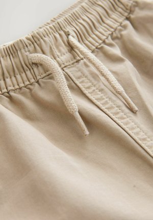 Pantaloni corti beige con vita elastica e tessuto arricciato, dotati di un cordoncino intrecciato per la regolazione. Texture liscia e dettagli di design minimi.