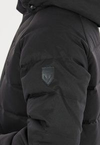 Doudoune noire avec un design matelassé, dotée d'un patch en cuir texturé sur le bras supérieur et d'une fermeture à bouton sécurisée.